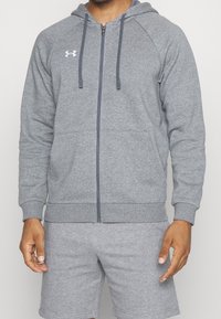 Sudadera gris con cremallera, bolsillo frontal, capucha ajustable y puños de canalé. Presenta un ligero logo en el pecho y está hecha de un material suave.