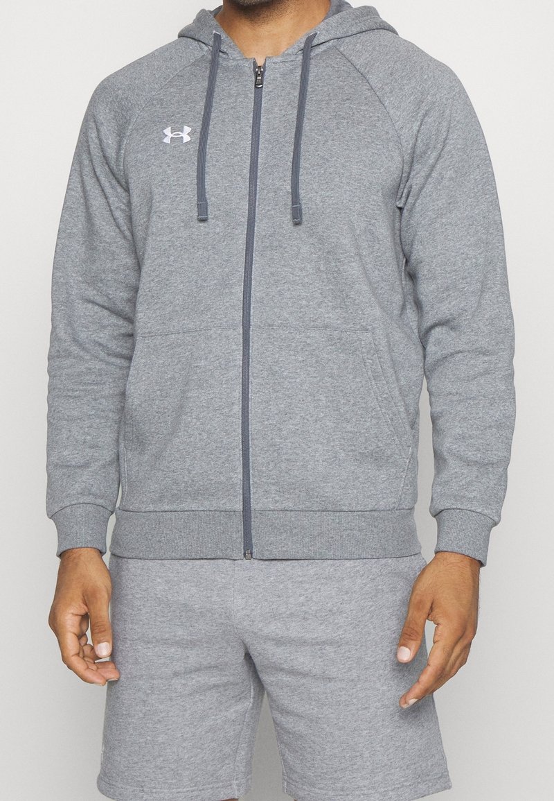 Sudadera gris con cremallera, bolsillo frontal, capucha ajustable y puños de canalé. Presenta un ligero logo en el pecho y está hecha de un material suave.