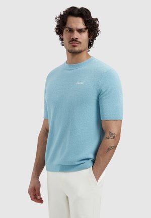 Man met krullend donker haar die een lichtblauwe korte mouw trui en witte broek draagt, staat met één hand in de zak tegen een neutrale achtergrond.