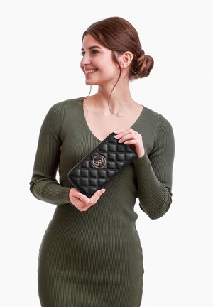 Black quilted leather wallet with a gold emblem, held by a person in a ribbed olive green dress against a white background.
