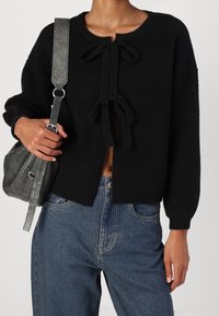 Cárdigan de punto negro con cuello redondo, que presenta dos lazos en la parte delantera, usado sobre jeans azules y combinado con un bolso texturizado gris.
