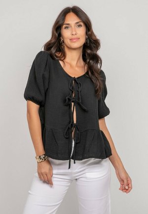 Femme aux longs cheveux ondulés portant un chemisier noir à manches bouffantes avec des liens à l'avant et un pantalon blanc, debout devant un fond clair uni.