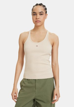 Donna con i capelli intrecciati che indossa una canotta beige a coste e pantaloni verde oliva, in piedi davanti a uno sfondo bianco semplice.