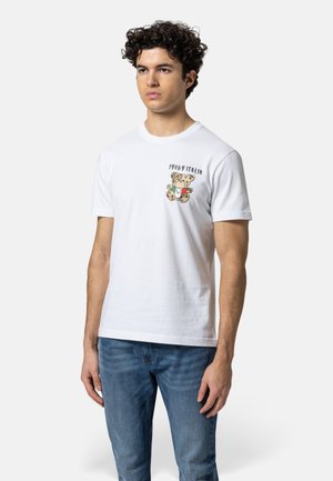 Junger Mann mit lockigem Haar trägt ein weißes T-Shirt mit einem kleinen Teddybär und dem Schriftzug "1969 ITALIA" auf der Brust, kombiniert mit blauen Jeans.