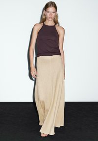Donna che indossa un top brown scuro senza maniche e una lunga gonna beige, in piedi contro uno sfondo bianco e nero.