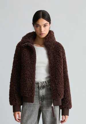 Femme aux cheveux foncés portant une veste marron texturée zippée, un haut blanc côtelé et un jean gris taille haute, debout devant un fond uni.