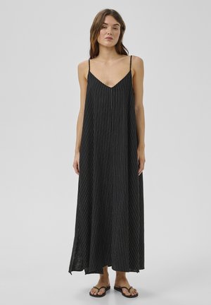 Žena v čiernych maxi šatách s tenkými bielymi vertikálnymi pruhmi a tenkými ramienkami, kombinovaných s čiernymi šľapkami, stojaca na jednoduchom pozadí.