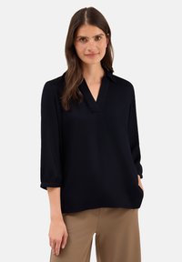 Frau mit langen braunen Haaren und Sommersprossen, die eine schwarze V-Ausschnitt-Bluse und beige Hosen trägt, vor einem schlichten weißen Hintergrund.