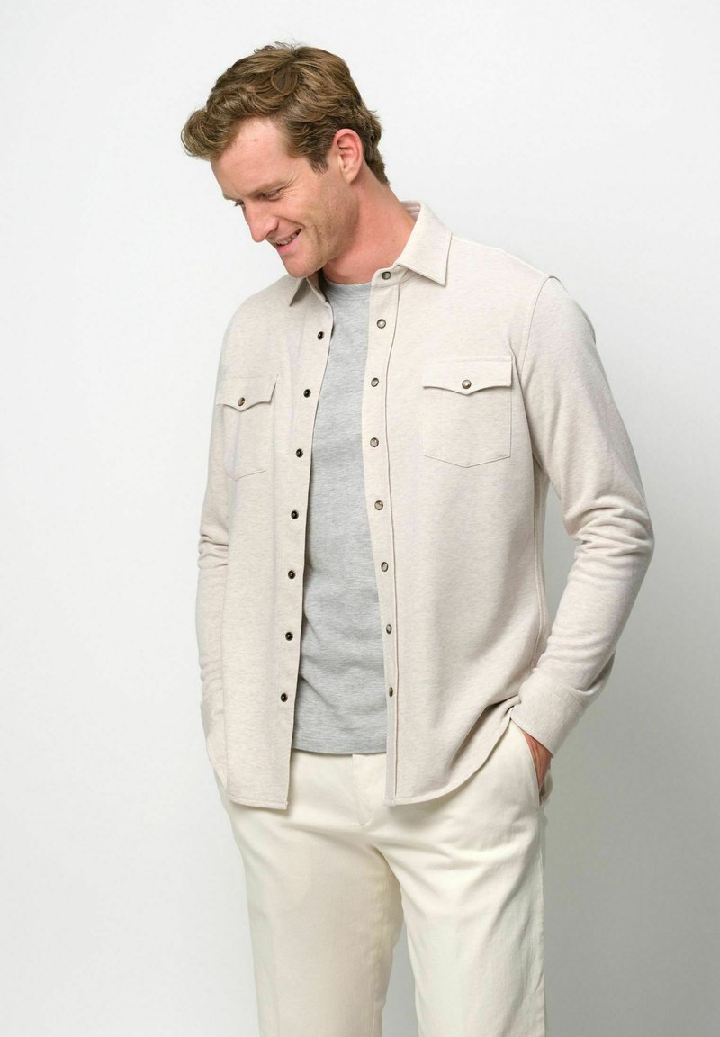 Chemise beige clair à boutons avec deux poches poitrine et boutons foncés, portée sur un t-shirt gris, assortie à un pantalon crème clair.