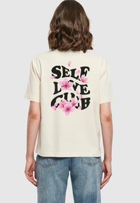 Krémové bavlnené tričko s čiernou grafikou "SELF LOVE CLUB" obklopenou ružovými kvetinovými akcentmi na chrbte. Pohodlný strih.