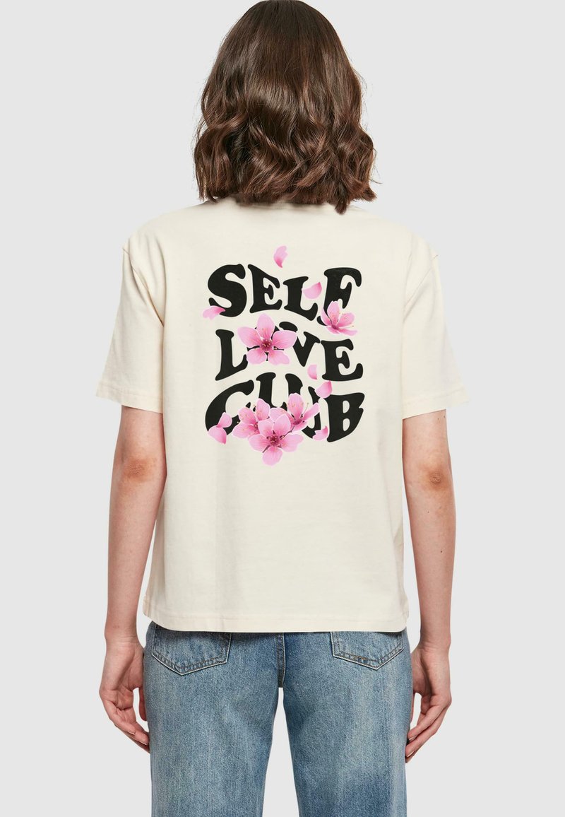 Krémové bavlněné tričko s černou grafikou "SELF LOVE CLUB" obklopenou růžovými květinovými akcenty na zadní straně. Uvolněný střih.