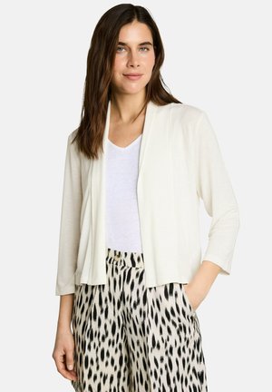 Woman with long brown hair wearing a white open cardigan, white top, and high-waisted black and white spotted pants, hands relaxed by sides.
