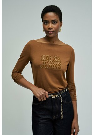 Femme portant un haut marron à manches longues avec le texte "SALSA JEANS", jean foncé avec ceinture chaîne dorée, debout devant un fond clair uni.