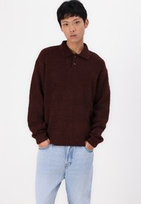Maglione in maglia marrone, con texture, colletto a polo e tre bottoni. Vestibilità ampia, polsini a coste, abbinato a jeans in denim blu chiaro.