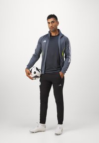 Grijze zip-up jas met witte en neon accenten, zwarte broek, houdt een zwart-wit voetbalt in de hand, draagt witte voetbalschoenen.