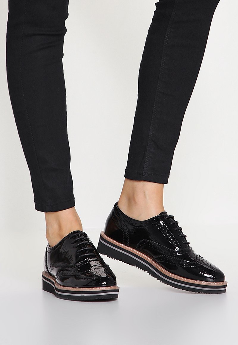Chaussures brogues en cuir verni noir avec semelles épaisses à rayures portées avec un pantalon noir slim sur un fond clair uni.