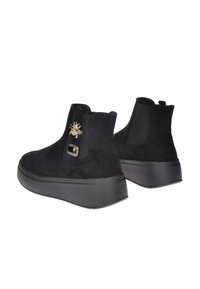 Stivaletti neri in suede con suole spesse, decorati con una spilla a forma di ape dorata e una gemma nera sul lato esterno.