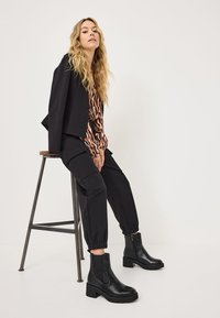 Black leather ankle boots with chunky soles, elastic side panels, paired with black trousers and a patterned blouse, seated on a metal stool.