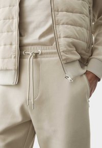 Beige quiltet vest over en lyse beige crewneck, parret med matchende tapered sweatpants med snøre og lynlåslukning ved anklerne.