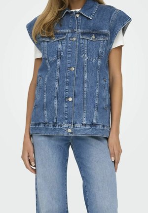 Mujer con chaleco sin mangas de mezclilla azul con cierre de botones sobre camisa blanca de manga corta y pantalones vaqueros de mezclilla azul claro.