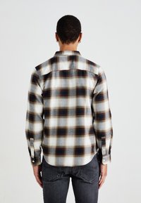 Camisa de franela a cuadros en tonos blanco, gris y rojo oscuro; cuenta con mangas largas, puños abotonados y un corte relajado con un dobladillo redondeado.