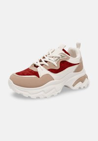 Les baskets présentent un mélange de matériaux beiges, rouges et blancs, avec une tige texturée, une semelle blanche épaisse et des accents métalliques argentés.