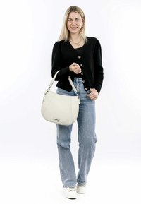 White leather shoulder bag with a textured finish, curved shape, and an adjustable strap, held by a person wearing a black cardigan and jeans.