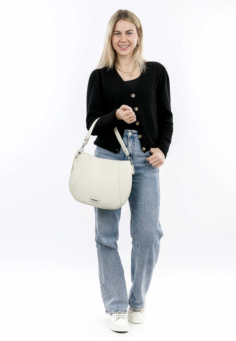 White leather shoulder bag with a textured finish, curved shape, and an adjustable strap, held by a person wearing a black cardigan and jeans.