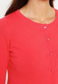 Korallfärgad cardigan i mjukt stickat tyg, med rund halsringning, sex matchande knappar fram och små öljett-detaljer längs kanten.