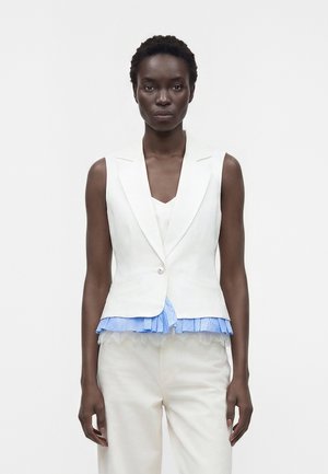 Femme portant un blazer blanc sans manches sur un chemisier bleu à volants et un pantalon blanc, debout devant un fond blanc uni.