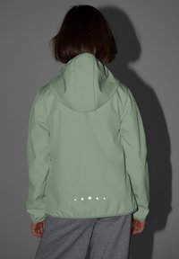 Personne portant une veste à capuche vert clair avec des points réfléchissants dans le dos, debout devant un fond gris.