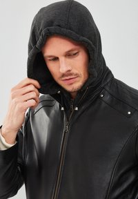 Veste en cuir noir avec fermeture éclair, dotée d'une capuche en polaire grise et de détails matelassés aux épaules. Texture lisse avec des accessoires en métal argenté.