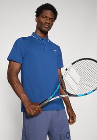 Plava polo majica s ravnim ovratnikom, dva gumba i svijetlim detaljima. Model drži tenisku reketa, nosi srebrne kratke hlače s logom.