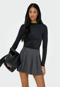 Kvinne med langt svart hår iført en svart rynket langermet topp, grå plisserte miniskjørt, holdende en svart lærveske i den ene hånden.