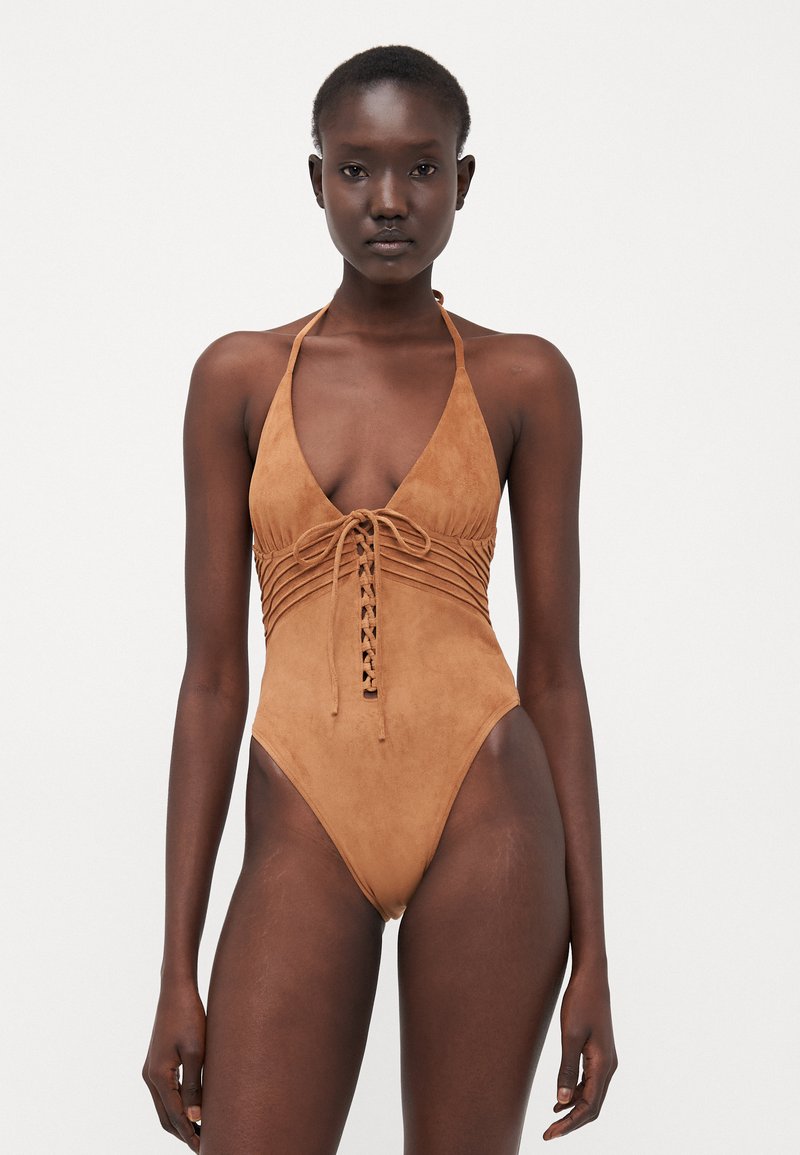 Brown suede one-piece swimsuit with a deep V-neck, front lacing detail, and halter neck straps. Smooth texture and minimalist design.