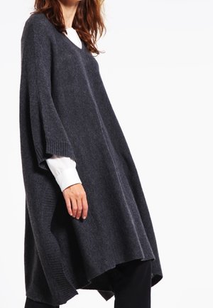 Frau trägt einen locker sitzenden, dunkelgrauen V-Ausschnitt Strickponcho über einem weißen Langarmshirt und schwarzen Hosen, steht vor einem weißen Hintergrund.