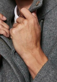 Manos ajustando el cuello de un abrigo de lana gris con cierre de cremallera y botón de presión.