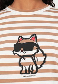 Pruhovaná béžová a biela košeľa s kreslenou mačkou, ktorá má čierne slnečné okuliare, a textom "KARL LAGERFELD" pod obrázkom mačky.