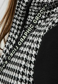 Schwarze und weiße Hahnentrittmuster-Überziehweste mit strukturiertem Stoff, schwarzem Körper und einem Markenband entlang des Reißverschlusses für Details.