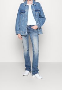 Veste en jean délavée clair avec deux poches poitrine et un col à revers, associée à un jean bleu foncé et des baskets blanches. Coupe décontractée.