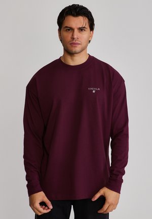 Mężczyzna ubrany w jednolity, ciemnofioletowy longsleeve z małym białym logo "SikSilk" na lewej piersi, stojący na neutralnym tle.