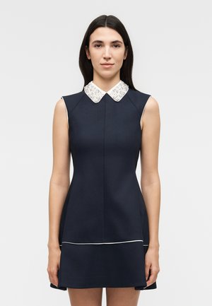 Γυναίκα με αμάνικο μπλε φόρεμα με λευκό κολάρο με χάντρες, που στέκεται μπροστά σε απλό ανοιχτό γκρι φόντο.