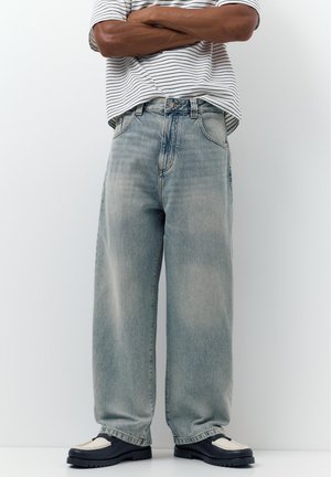 Osoba oblečená v svetlomodrých džínsach s širokými nohavicami, čierno-bielych mokasínach a bielej pruhovanej košeli so založenými rukami stojaca opretá o stenu.