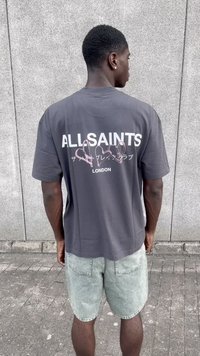 Camiseta gris de gran tamaño con gráficos de "ALLSAINTS" y un corazón rosa en la parte de atrás, combinada con pantalones cortos de mezclilla claros y una textura de color sólido.
