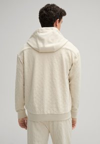 Felpa beige con cappuccio caratterizzata da un pattern superficiale testurizzato, tasca a marsupio e cappuccio con cordino regolabile. Polsini e vita aderenti.