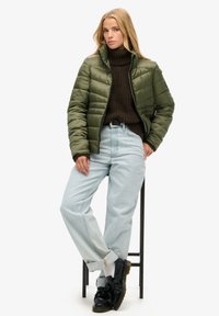Oliwkowa kurtka puffer z poziomymi przeszyciami, brązowy sweter z dzianiny, jasnoniebieskie dżinsy oraz czarne buty ze sznurówkami. Teksturowany materiał.
