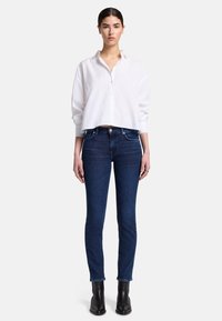 Weißes, cropped Hemd mit Knöpfen, kombiniert mit dunkelblauen Skinny Jeans. Schwarze Stiefeletten runden das Outfit ab. Saubere Linien und eine figurbetonte Silhouette.