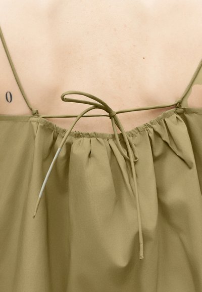 Οπίσθια όψη ενός ατόμου που φοράει τοπ ελιάς με λεπτές τιράντες δεμένες με φιόγκο στο άνω μέρος της πλάτης, αποκαλύπτοντας ένα μικρό τατουάζ κοντά στον ώμο.