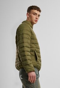Olivgrön puffjacka med hög krage, horisontell sömnad, sidofickor och slät yta, bärs över texturerade jeansbyxor.