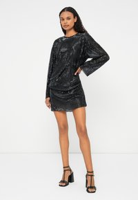 Melnas strūklas kleita ar garām piedurknēm, strukturēta auduma un drapēta dizaina. Apvienota ar melniem siksnu zābakiem. Gluda, spīdīga virsma.